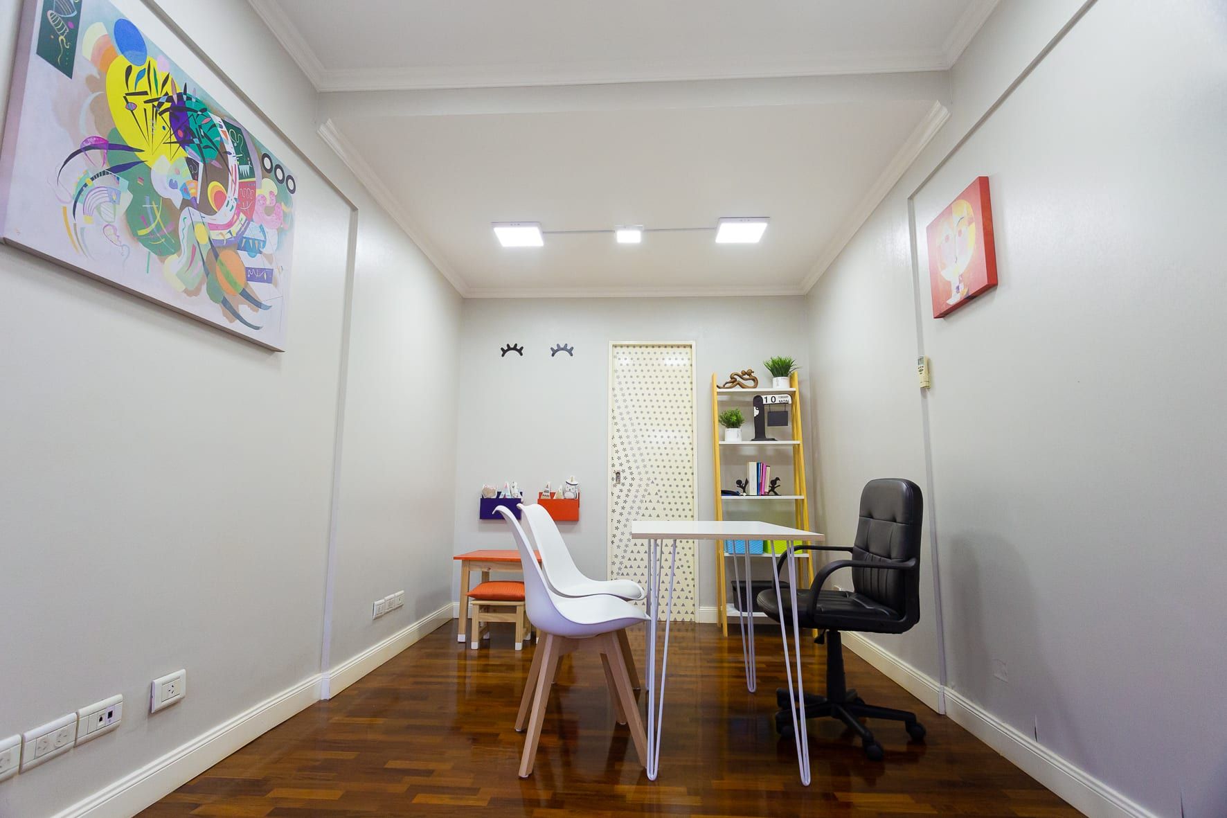 Ambiente con piso de madera lustrada equipado como consultorio: escritorio, sillas y rincón infantil con mesa naranja
