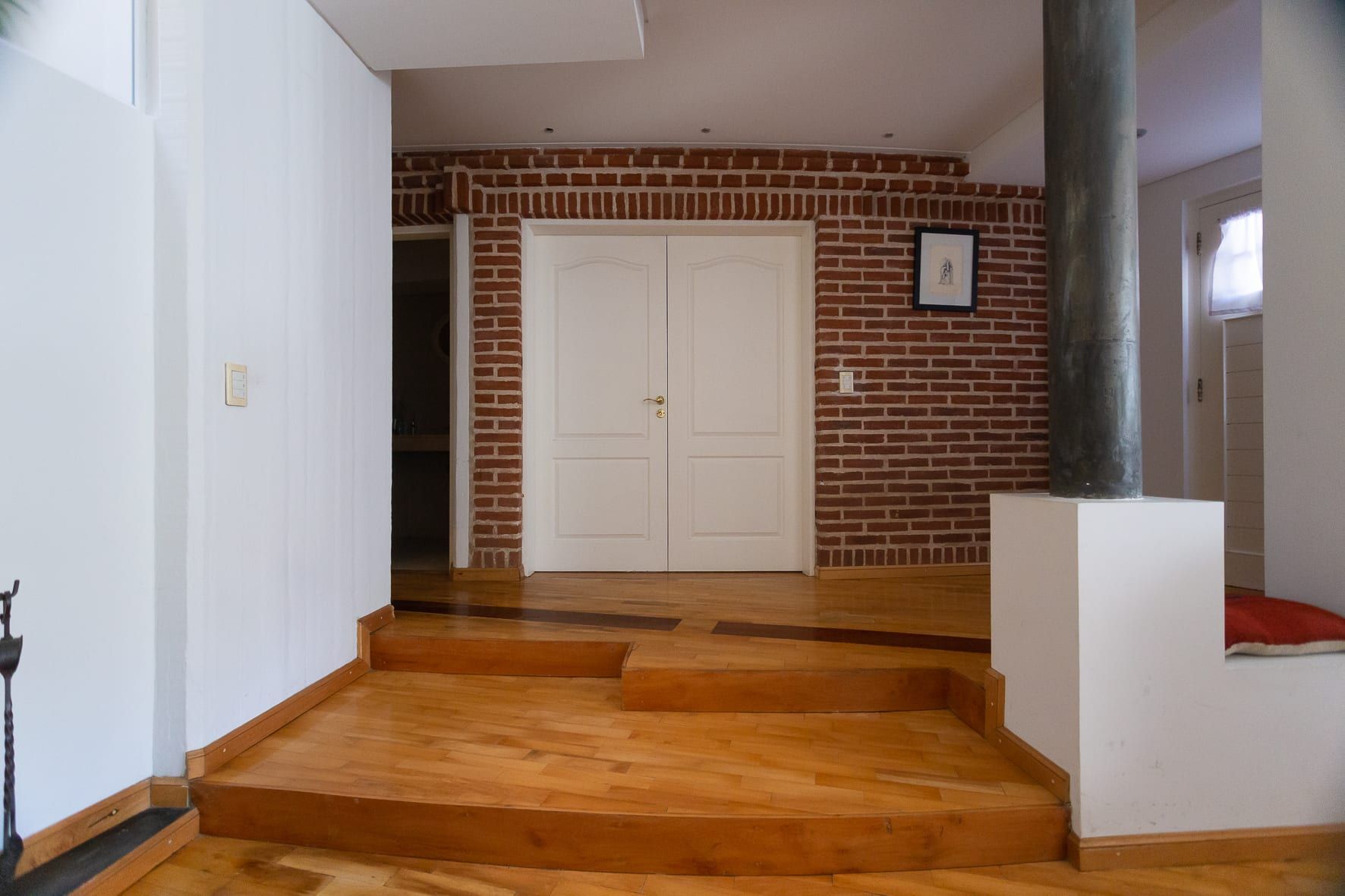 Hall con muro de ladrillo visto, puerta doble blanca y desniveles de madera en la casa Chiclana