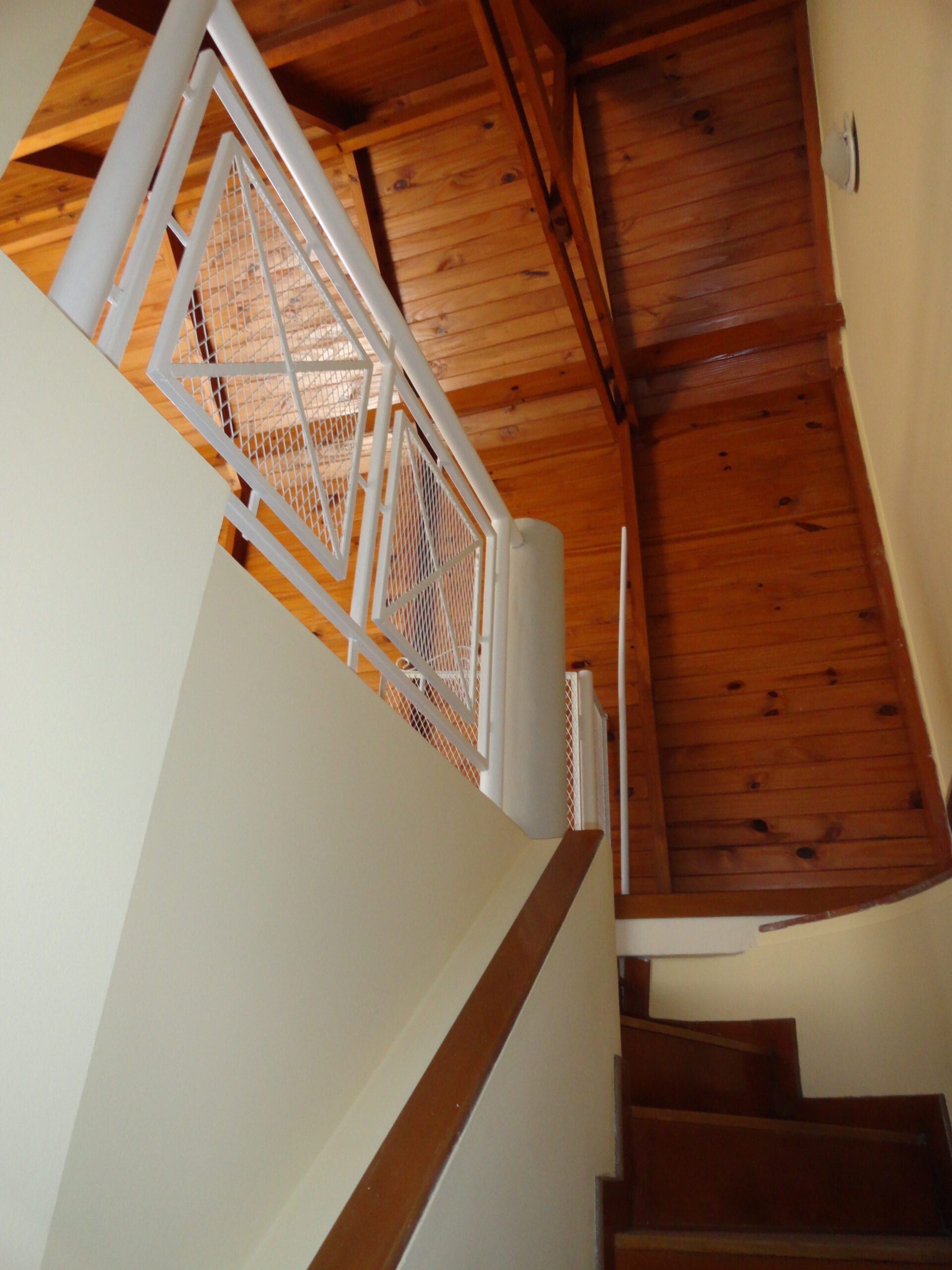 Escalera de madera con barandas metálicas blancas que sube al entrepiso, bajo el cielorraso de pino a la vista