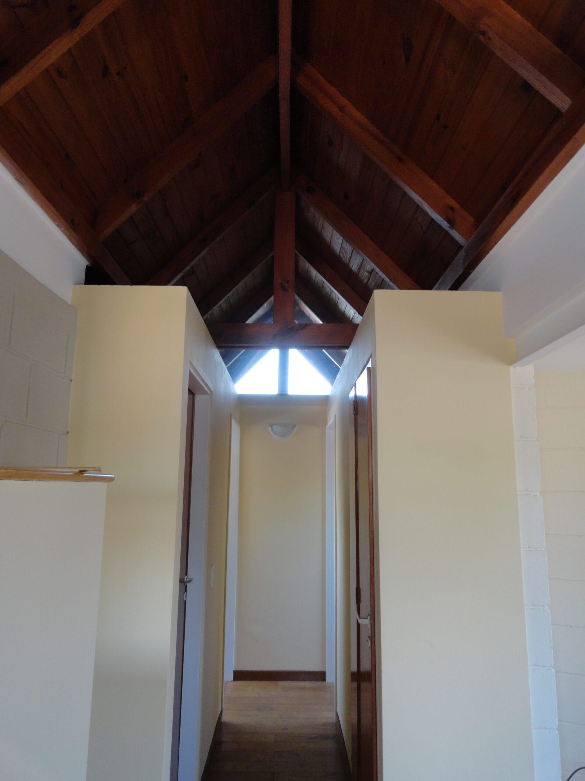 Pasillo de dormitorios con puertas de madera, coronado por el techo de pino a dos aguas y ventana triangular en la cumbrera
