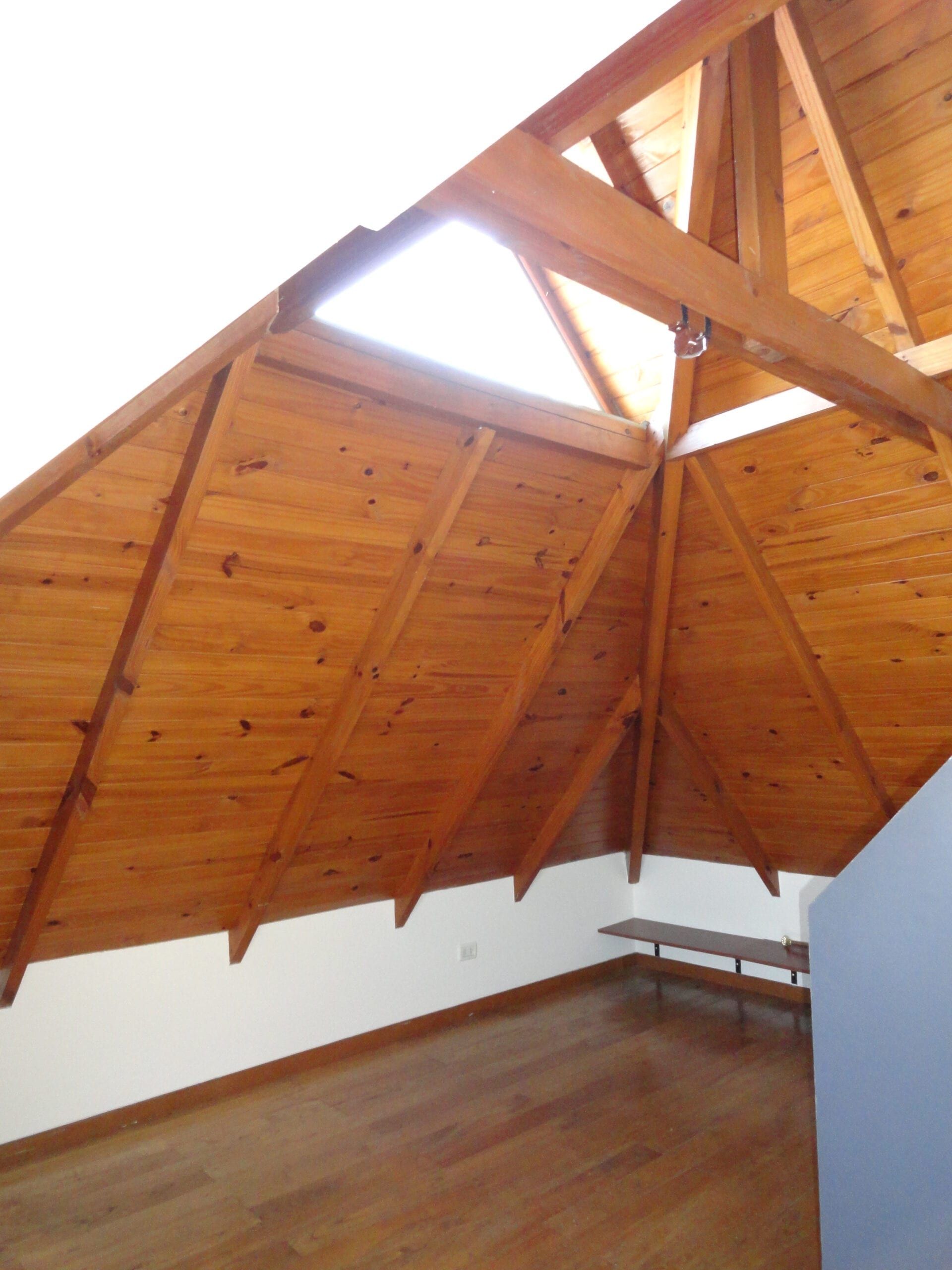 Dormitorio en planta alta bajo la pendiente del techo de madera, con lucarna que aporta luz natural y piso de madera